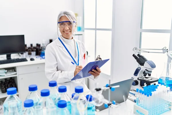 Orta yaşlı, gri saçlı, bilim adamı üniforması giyen, laboratuarda çalışan bir kadın.