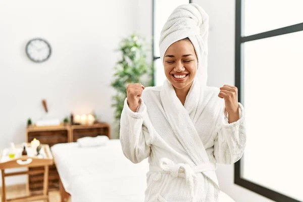 Young brunette woman wearing towel and bathrobe standing at beauty center excited for success with arms raised and eyes closed celebrating victory smiling. winner concept.