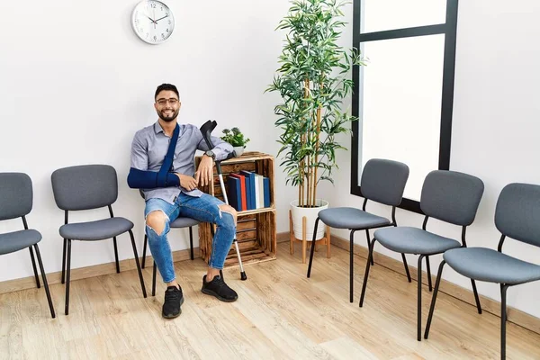 Young arab man smiling confident sitting on chair with arm sling holding crutch at clinic waiting room