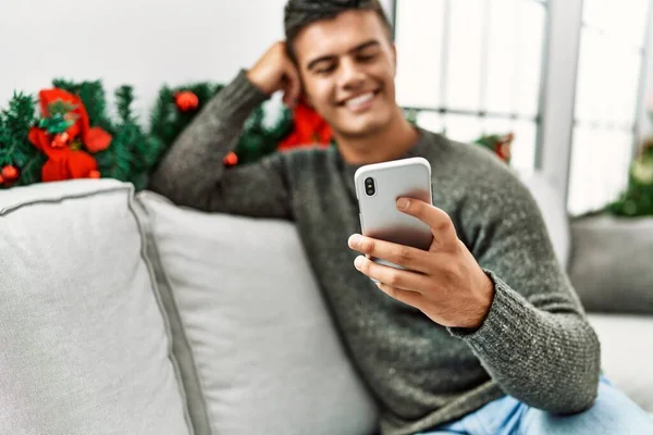 Young hispanic man using smartphone sitting on sofa by christmas tree at home