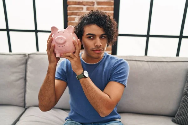 Young Hispanic Man Holding Piggy Bank Sitting Sofa Home — Zdjęcie stockowe