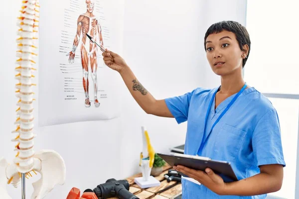 Young hispanic woman wearing physiotherapist uniform pointing to vertebral column holding checklist at physiotherapy clinic