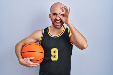 Sakallı, basketbol forması giyen, top tutan, el hareketleriyle gülümseyen, mutlu yüzüyle parmaklarının arasından bakan kel bir adam.. 