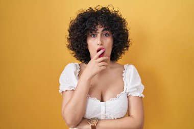 Sarı arka planda duran kıvırcık saçlı genç esmer kadın elleri çenesinde olan inançsızlıktan, şaşkınlıktan ve şaşkınlıktan büyülenmiş görünüyor. 