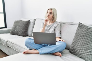 Young caucasian woman using laptop at home sitting on the sofa with hand on chin thinking about question, pensive expression. smiling with thoughtful face. doubt concept. 