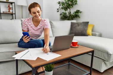 Akıllı telefon ve dizüstü bilgisayar kullanan genç bir kadın evdeki kanepede oturuyor.