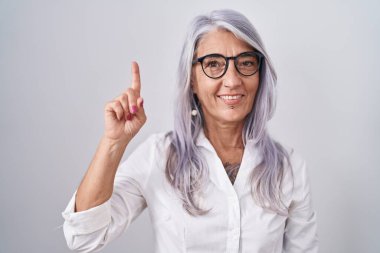 Beyaz arka planda duran gözlüklü orta yaşlı bir kadın bir numaralı parmağı gösterirken kendine güvenen ve mutlu bir şekilde gülümsüyor.. 