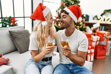 Mutlu bir şekilde gülümseyen genç çift Noel şapkası takıyor evde şampanyayla kadeh kaldırıyor.