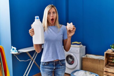 Beautiful woman doing laundry holding detergent bottle and piggy bank in shock face, looking skeptical and sarcastic, surprised with open mouth 