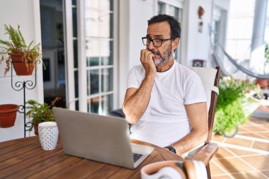 Middle age man using computer laptop at home looking stressed and nervous with hands on mouth biting nails. anxiety problem. 