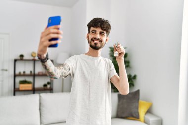 Genç İspanyol adam, elinde yeni evin anahtarı olan akıllı telefonun yanında selfie çekiyor.