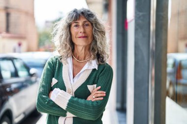 Orta yaşlı kadın gülümsüyor. Kollarını kavuşturup caddede duruyor.