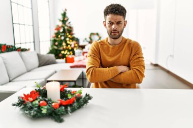 Arap genç adam Noel ağacının yanında masada oturuyor şüpheci ve gergin, yüzünde çapraz kollarla onaylamayan bir ifade var. negatif kişi. 