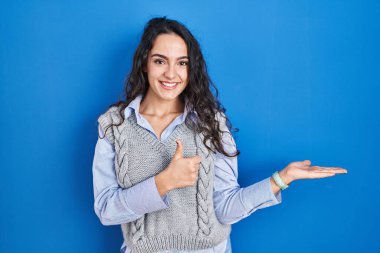 Mavi arka planda duran genç esmer kadın avuç içini gösteriyor ve baş parmaklarıyla iyi bir hareket yapıyor, mutlu ve neşeli gülümsüyor. 