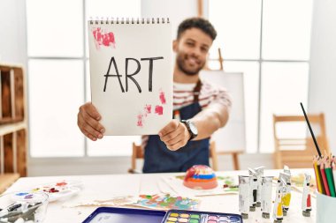 Genç Arap sanatçı gülümsüyor. Sanat stüdyosunda elinde sanat sözcüğüyle mutlu bir not defteri tutuyor..