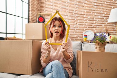 Yeni evindeki kanepede oturan genç beyaz kadın çılgınlığı kutluyor ve başarıya hayret ediyor gözleri açık heyecanla bağırıyor. 