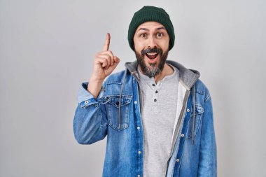 Young hispanic man with tattoos wearing wool cap pointing finger up with successful idea. exited and happy. number one. 