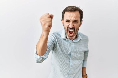 Middle age hispanic man with beard standing over isolated background angry and mad raising fist frustrated and furious while shouting with anger. rage and aggressive concept. 