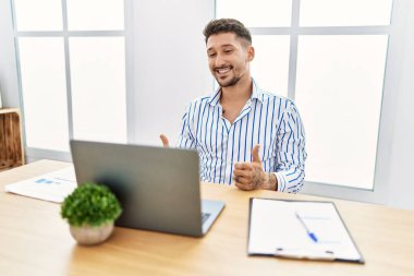 Ofiste çalışan sakallı yakışıklı genç adam bilgisayardaki başarı işaretini kullanıyor. Elleriyle olumlu jest yapıyor. Gülümsüyor ve mutlu. Neşeli ifade ve kazanan jest. 