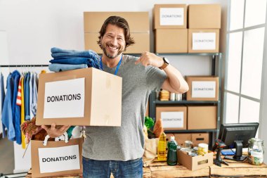 Handsome middle age man holding donations box for charity at volunteer stand looking confident with smile on face, pointing oneself with fingers proud and happy. 