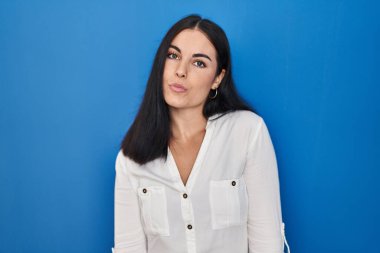 Genç İspanyol kadın mavi arka planda dikilip kameraya bakıyor ve güzel ve seksi bir şekilde yayına öpücük yolluyor. aşk ifadesi. 