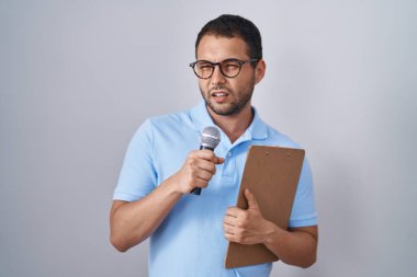 Hispanic man holding reporter microphone and clipboard clueless and confused expression. doubt concept. 
