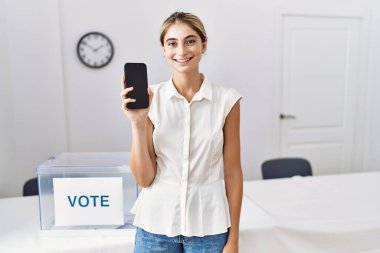 Siyasi seçimde elinde akıllı telefon tutan genç sarışın kadın pozitif ve mutlu görünüyor ve kendine güvenen bir gülümsemeyle dişlerini gösteriyor. 