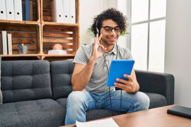 Kıvırcık saçlı İspanyol adam muayenehanede online seans yapıyor. Parmaklarıyla imza atıyor, gülümsüyor. Mükemmel bir sembol. 