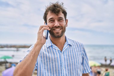 Genç İspanyol gülümseyen adam sahilde akıllı telefondan konuşarak mutlu oluyor.
