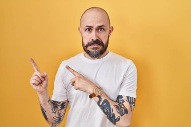 Young hispanic man with tattoos standing over yellow background pointing aside worried and nervous with both hands, concerned and surprised expression 