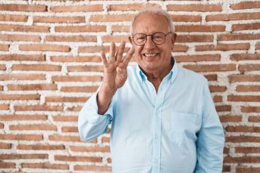 Senior man with grey hair standing over bricks wall showing and pointing up with fingers number four while smiling confident and happy. 