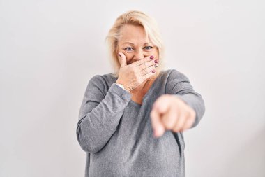 Beyaz arka planda dikilen orta yaşlı beyaz kadın sana gülüyor, eli ağzında kamerayı işaret ediyor, utanç ifadesi var. 