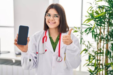 Genç İspanyol doktor kadın akıllı telefon ekranı gösteriyor mutlu ve pozitif gülümsüyor, başparmağı mükemmel ve onay işareti yapıyor. 