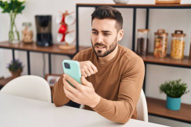 Genç İspanyol adam akıllı telefon kullanıyor. Evde masada oturuyor.
