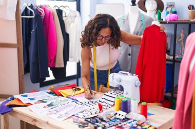 Middle age woman tailor holding t shirt writing on notebook at clothing factory