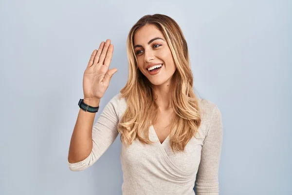 Young blonde woman standing over isolated background waiving saying hello happy and smiling, friendly welcome gesture