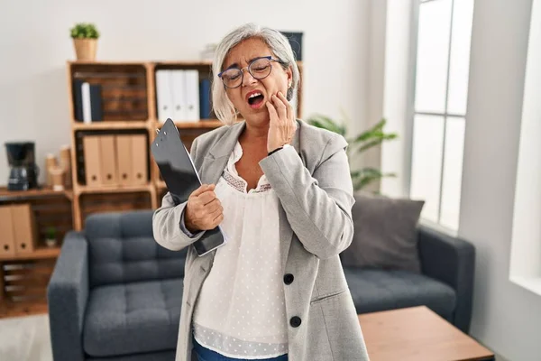 Middle age woman with grey hair at consultation office touching mouth with hand with painful expression because of toothache or dental illness on teeth. dentist concept.