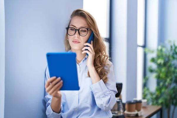 若いブロンドの女性のビジネスワーカーがオフィスでタッチパッドを使用してスマートフォンで話しています — ストック写真