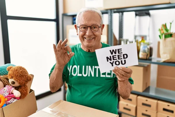 Senior volunteer man holding we need you banner doing ok sign with fingers, smiling friendly gesturing excellent symbol