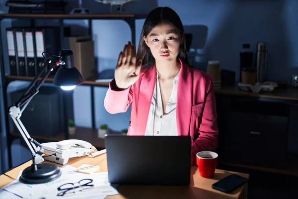 Chinese young woman working at the office at night doing stop sing with palm of the hand. warning expression with negative and serious gesture on the face.