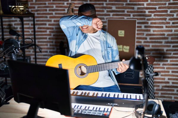 Hispanic young man playing classic guitar at music studio smiling cheerful playing peek a boo with hands showing face. surprised and exited 
