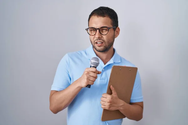 Hispanic man holding reporter microphone and clipboard clueless and confused expression. doubt concept.