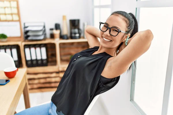 Young latin woman smiling confident relaxed with hands on head at office