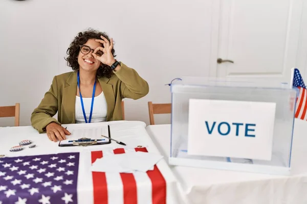 Young hispanic woman at political election sitting by ballot doing ok gesture with hand smiling, eye looking through fingers with happy face.