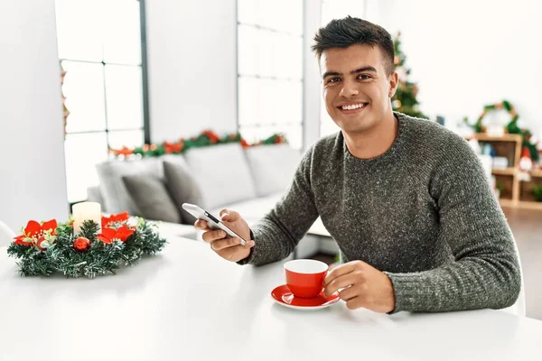 Young Hispanic Man Drinking Coffee Using Smartphone Sitting Christmas Tree — Stockfoto