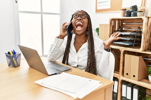 Black woman with braids working at the office speaking on the phone crazy and mad shouting and yelling with aggressive expression and arms raised. frustration concept.