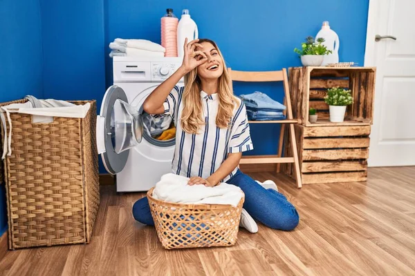 Young blonde woman doing laundry at home smiling happy doing ok sign with hand on eye looking through fingers