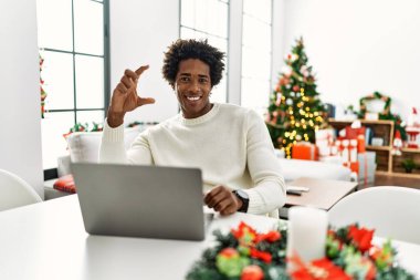 Genç Afro-Amerikalı adam dizüstü bilgisayar kullanıyor. Noel ağacının yanında oturuyor. Gülümsüyor ve el hareketleriyle küçük boy işareti yapıyor. ölçüm kavramı. 