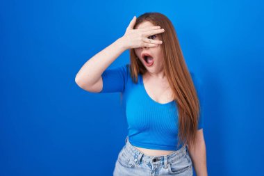 Kızıl saçlı kadın mavi arka planda dikiliyor. Şoka girip yüzünü ve gözlerini eliyle kapatıyor. Utanmış bir ifadeyle parmaklarından bakıyor.. 