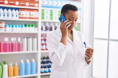 Afro-Amerikalı kadın eczacı elinde ilaç şişesiyle eczanede akıllı telefondan konuşuyor.